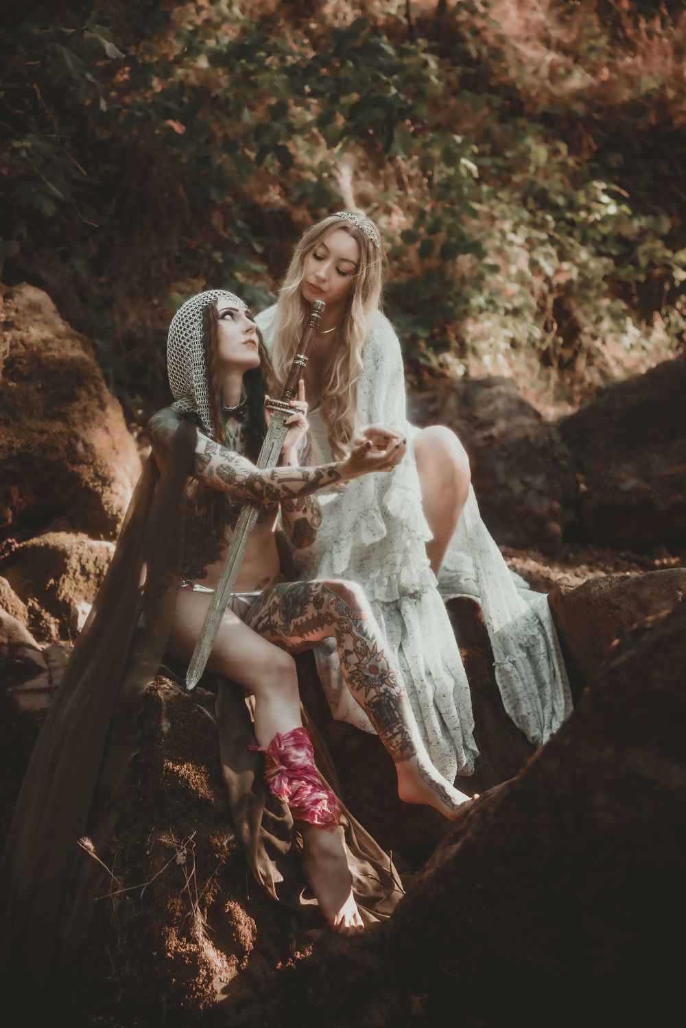 Two women in bohemian fantasy costumes sit among rocks in a forest, one in armor, one in a white lace gown.