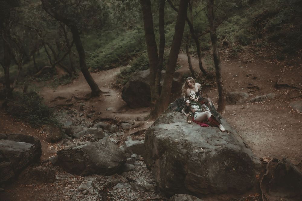 A couple sits intimately on a large rock surrounded by trees and a rocky stream in a moody forest setting.