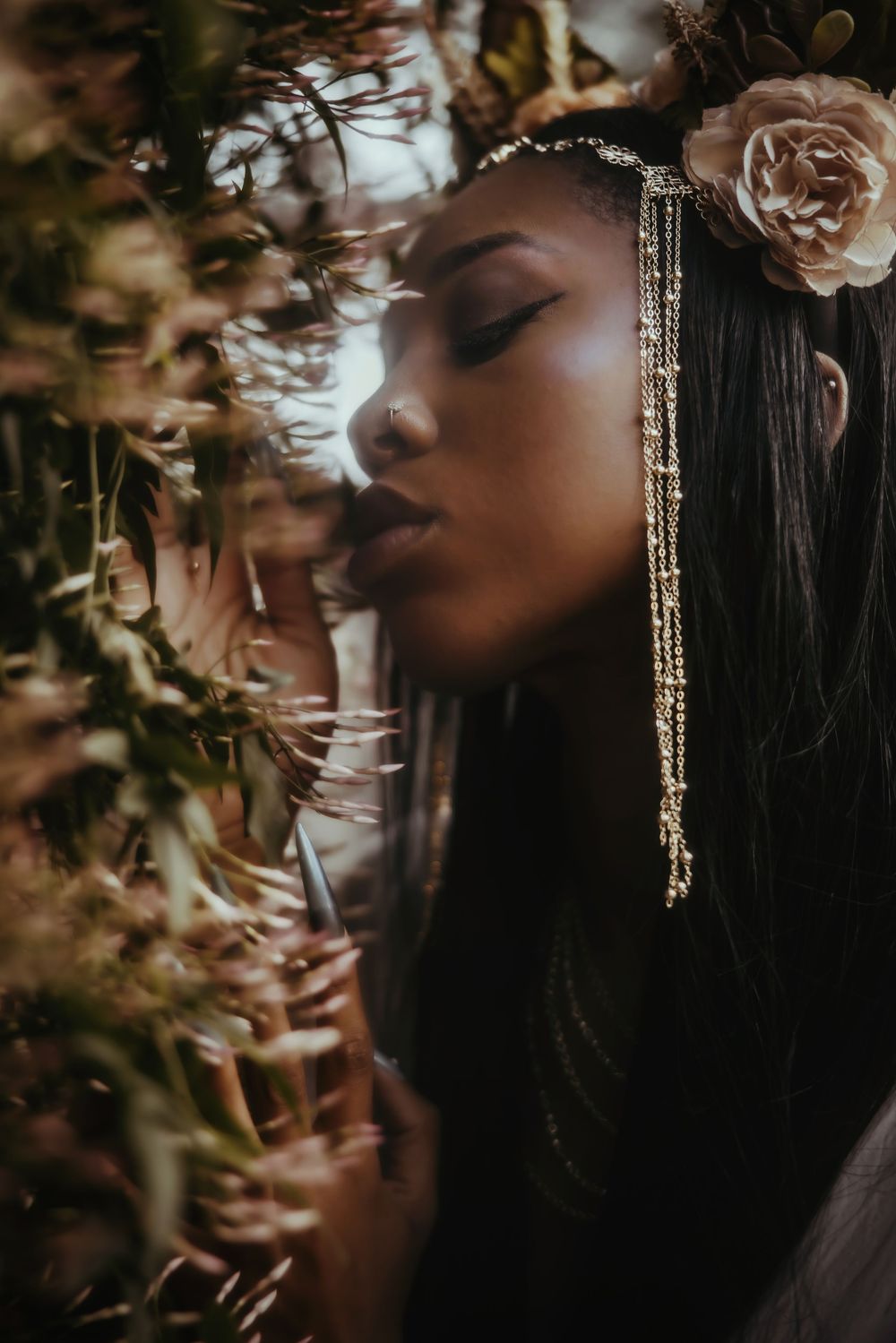 Artistic portrait of person with closed eyes wearing delicate gold jewelry and rose hair accessory among autumn foliage.