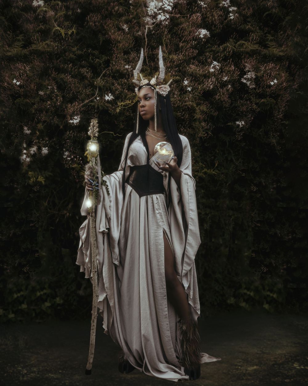 A mystical figure in flowing white robes stands in a dark garden holding a staff with glowing orbs and wearing antlers.