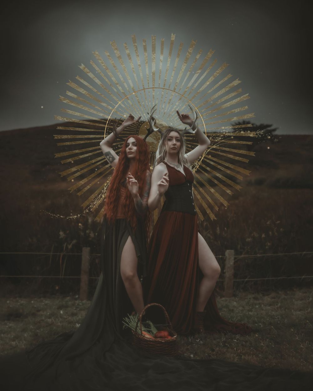 Two women in dark fantasy gowns pose before a golden sunburst halo with antlers, holding a harvest basket outdoors.