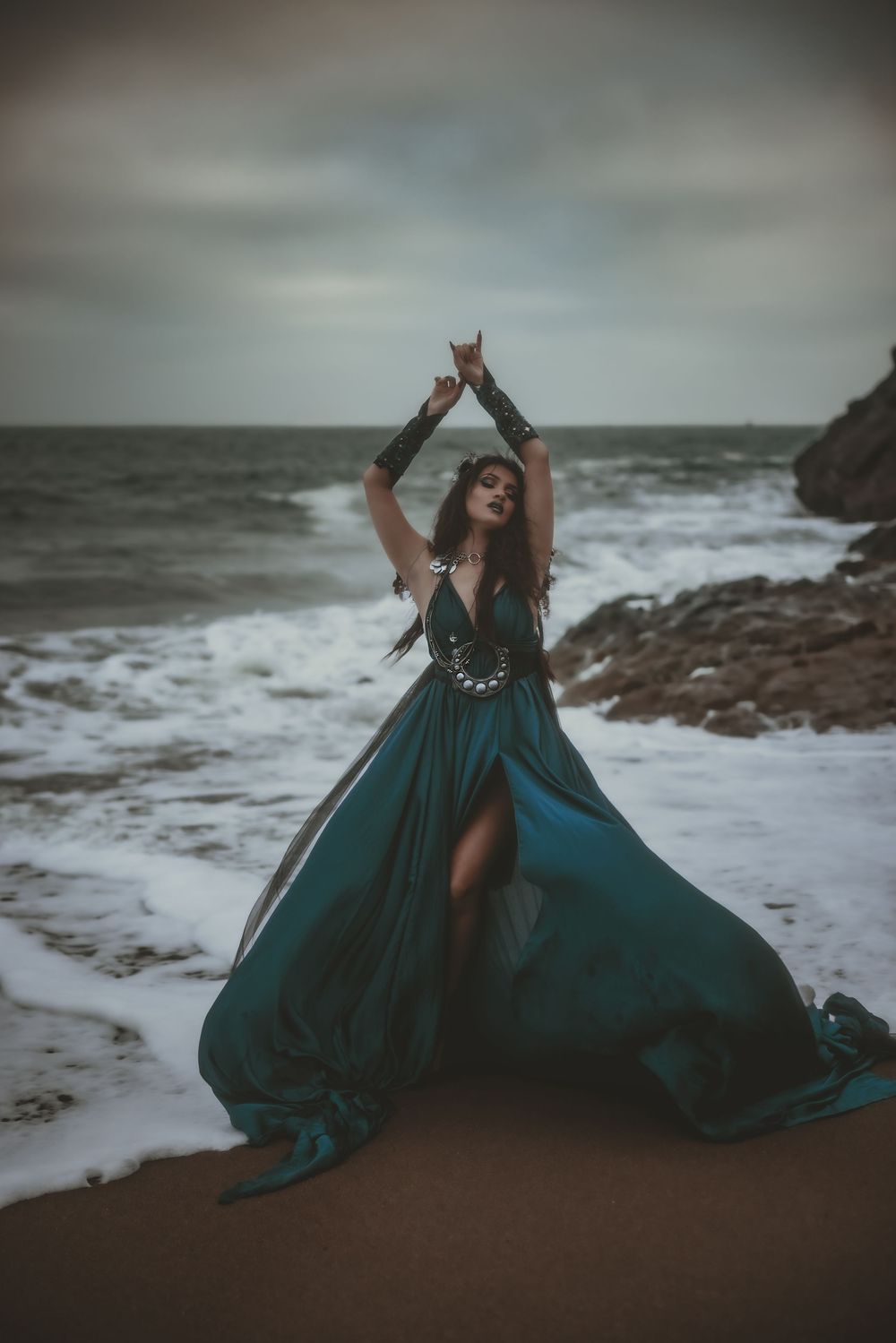 Woman in flowing teal gown poses dramatically on rocky beach with arms raised, waves crashing behind her.