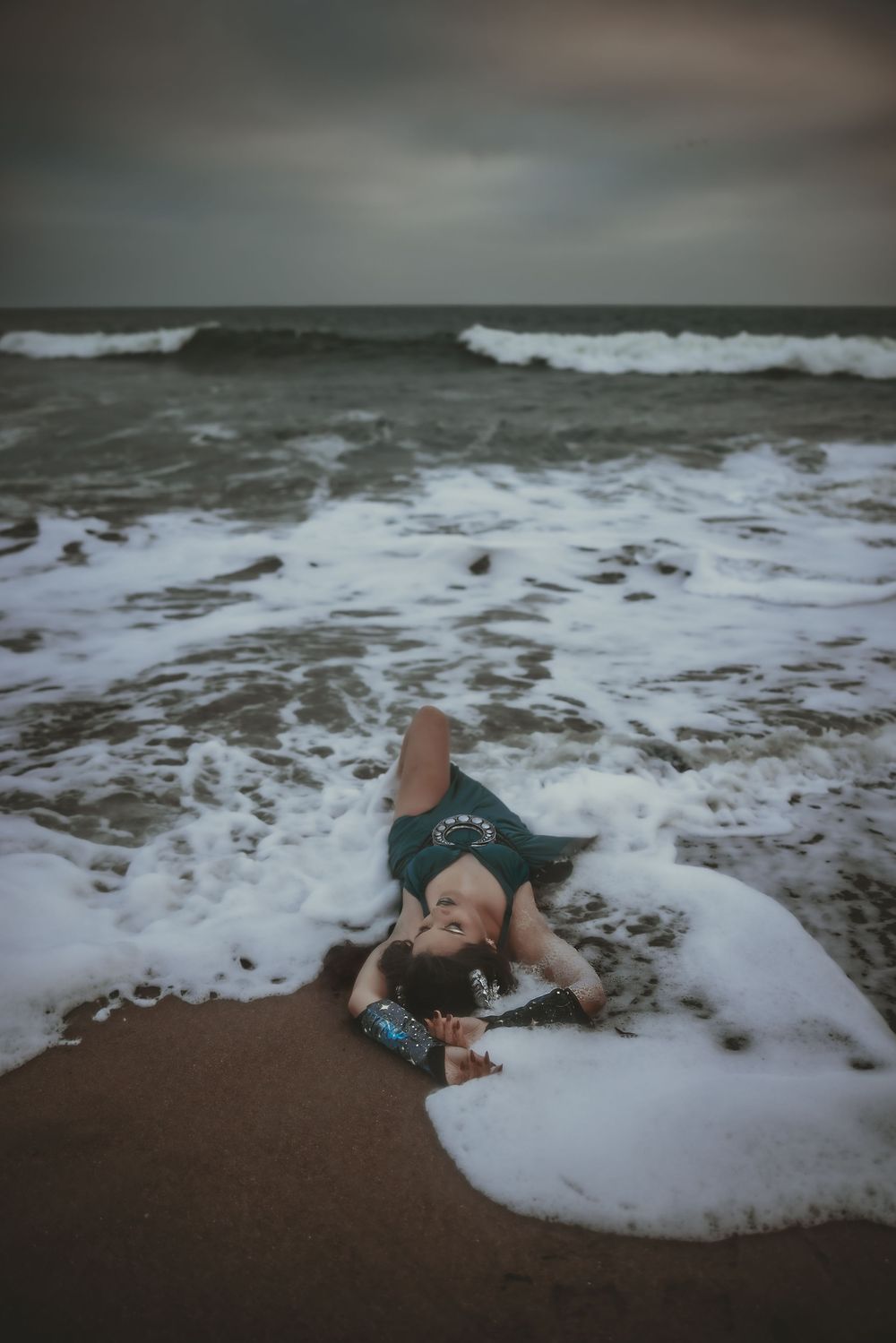 Woman in teal dress lying on sandy beach as ocean waves wash over her under dramatic stormy sky.