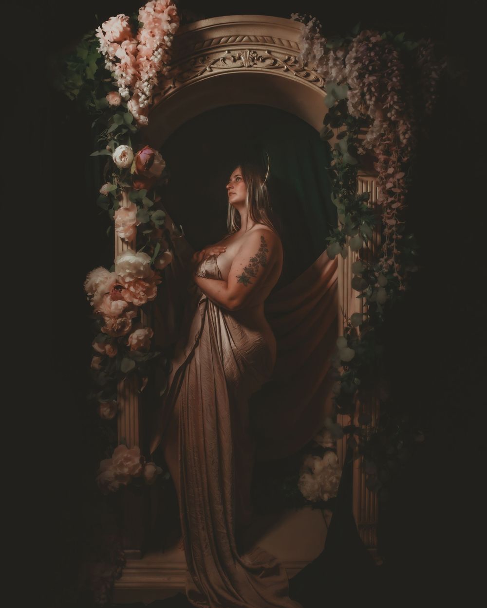 A woman in a flowing draped gown stands beneath a floral arch with roses, tattoos visible, in dramatic dark lighting.