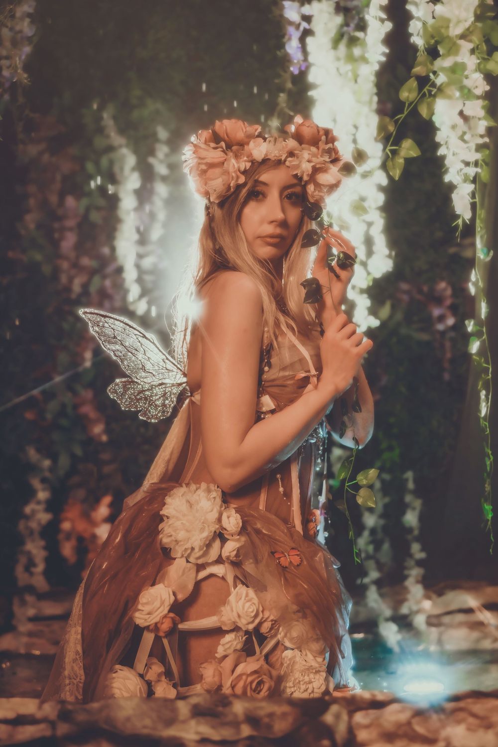 Woman dressed as a fairy in a floral gown and flower crown with glitter wings in an enchanted forest setting.