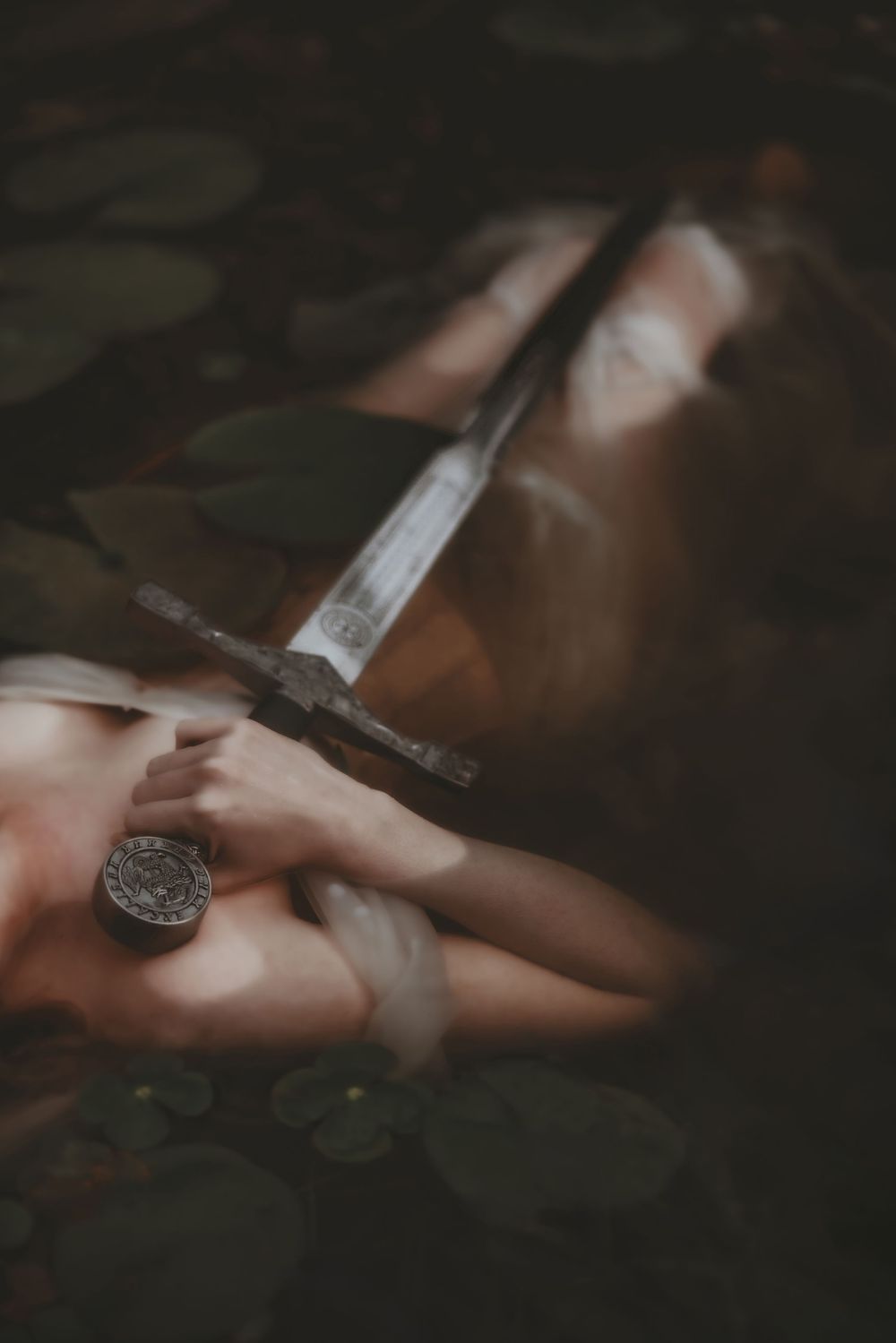 A person lying in dark water holding a medieval sword with lily pads, evoking an Arthurian legend scene.