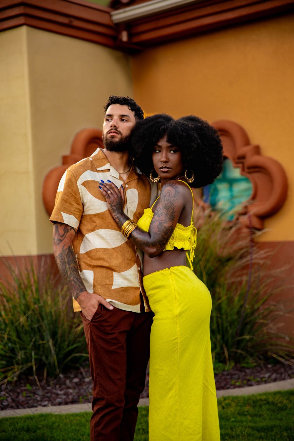 Stylish couple poses outdoors in vibrant outfits - brown patterned shirt and yellow dress against warm-toned building. best photographer in