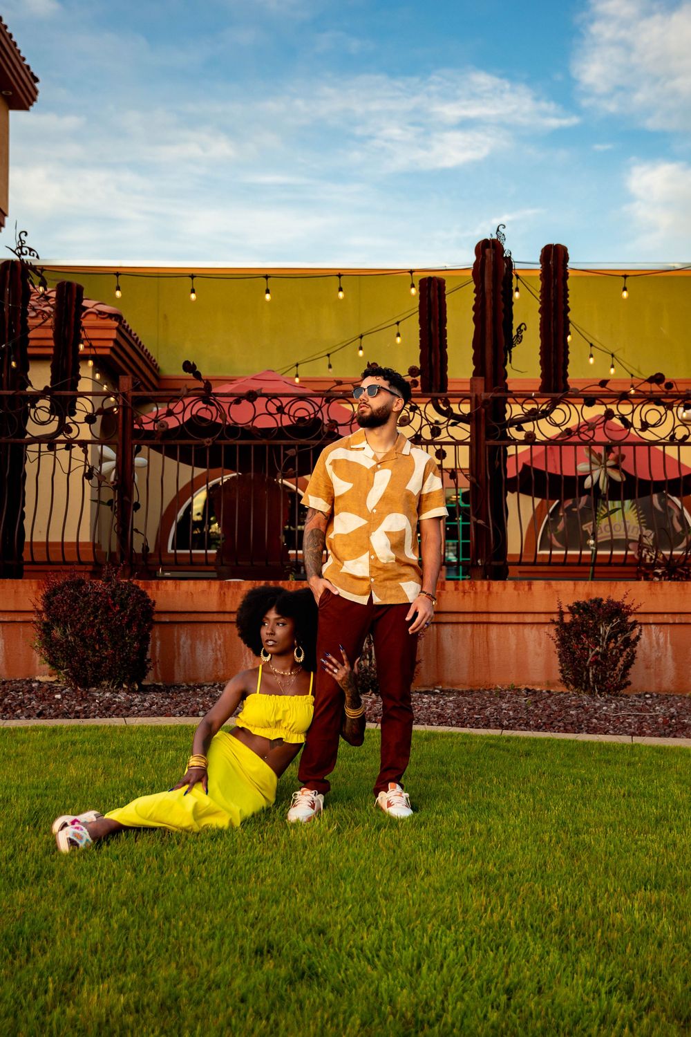 Stylish couple in vibrant yellow and brown outfits pose on grass with string lights and decorative railing in background.  best photographer