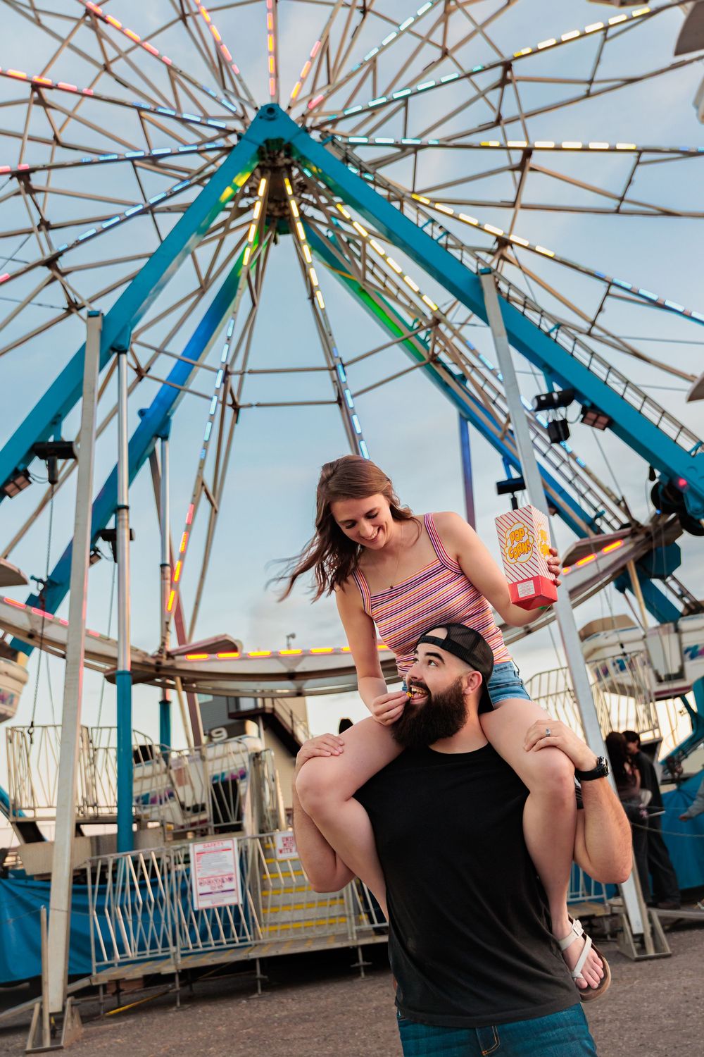 Couple at amusement park, woman sitting on man's shoulders holding popcorn, colorful Ferris wheel in background.