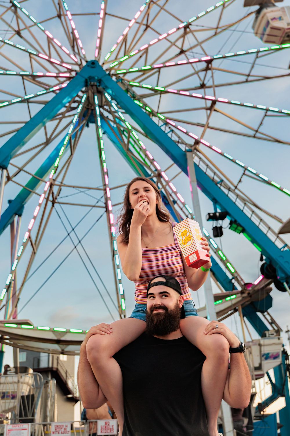 Couple at amusement park, woman sitting on mans shoulders eating popcorn in front of a colorful Ferris wheel.