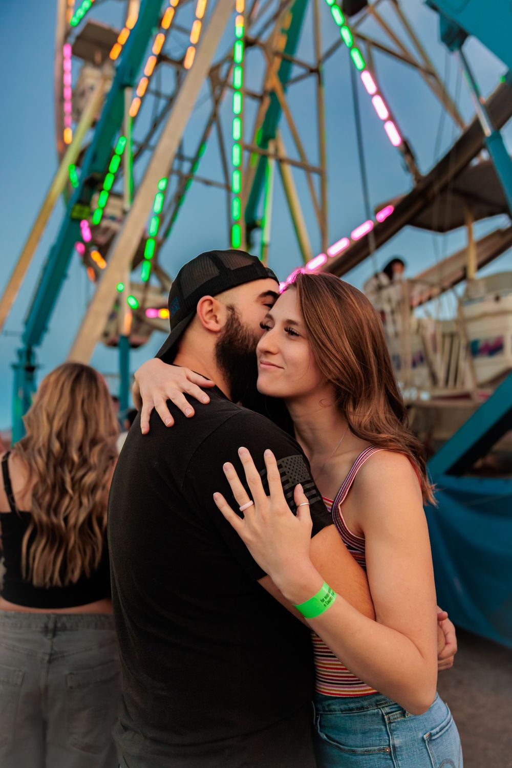 Romantic couple embracing and hugging in front of a colorful illuminated Ferris wheel at an amusement park.