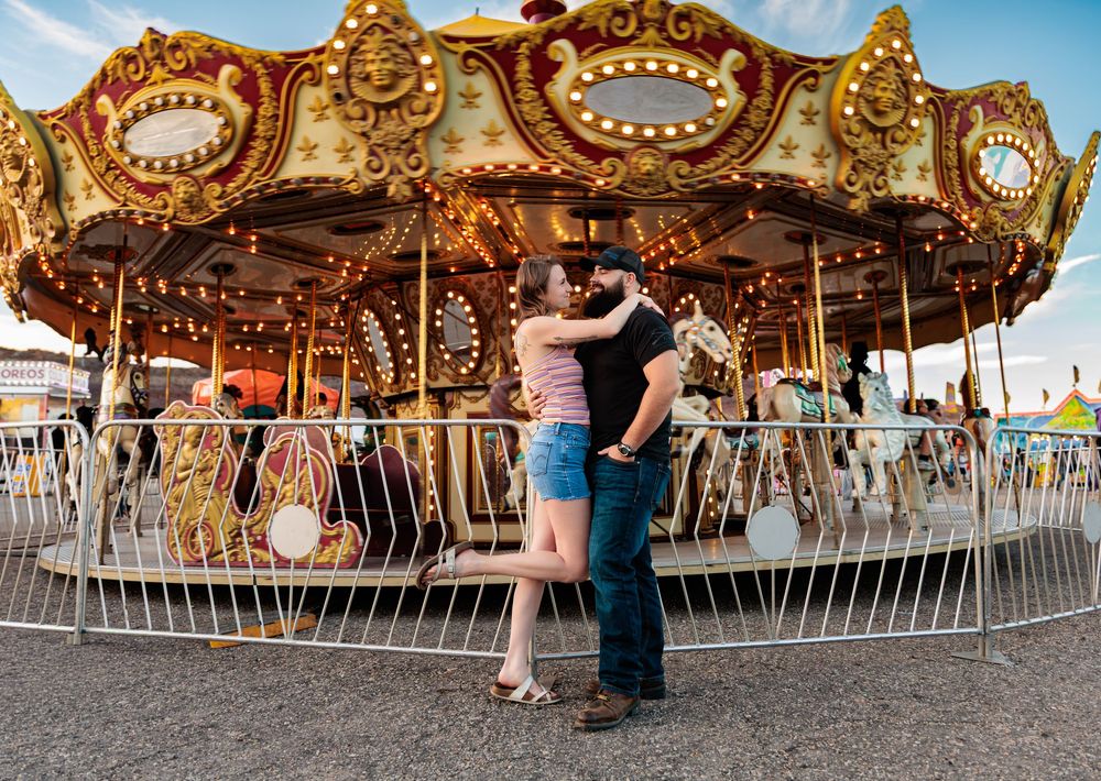 Couple sharing a romantic kiss in front of a colorful lit carousel at a fairground.