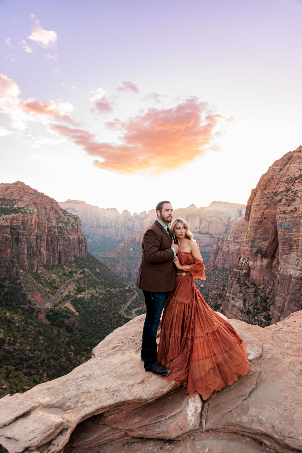 Couple embracing on a rocky overlook at Zion National Park during sunset, woman in flowing rust gown.