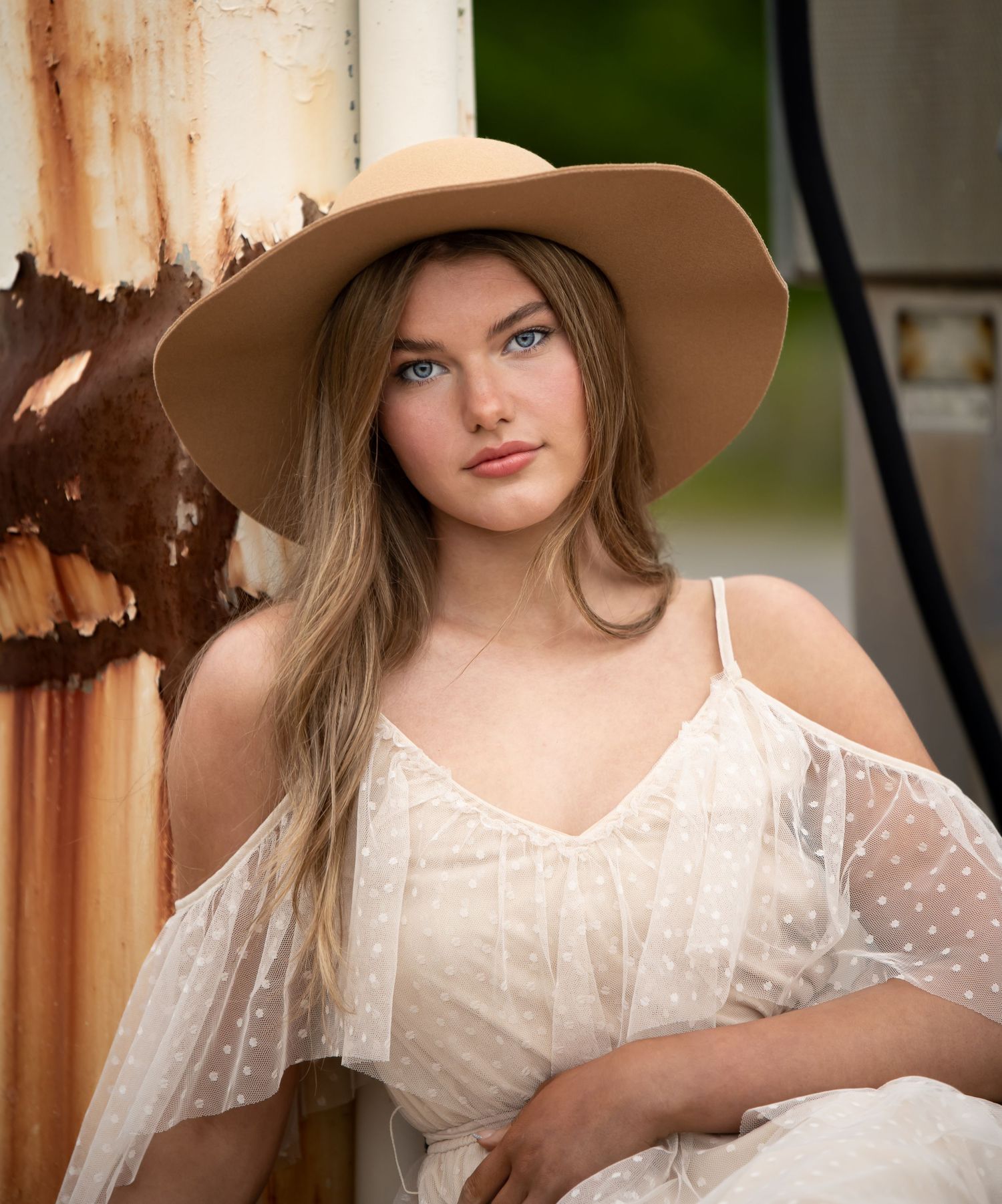 Portrait in ethereal white lace dress and wide brim hat against rustic background.