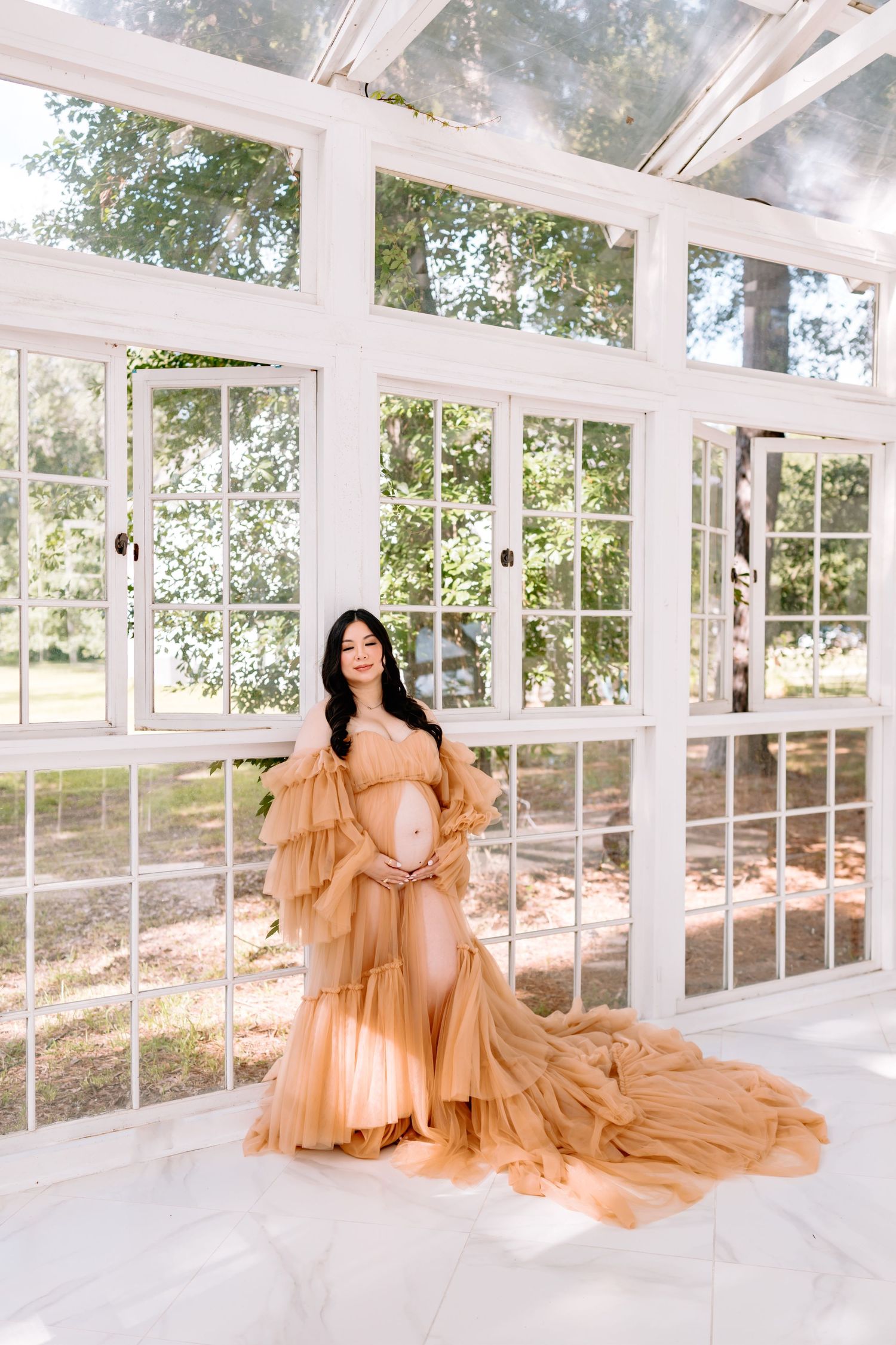 Pregnant mother in flowing peach gown during an elegant maternity photography session inside a glass house studio in Magnolia, Texas.