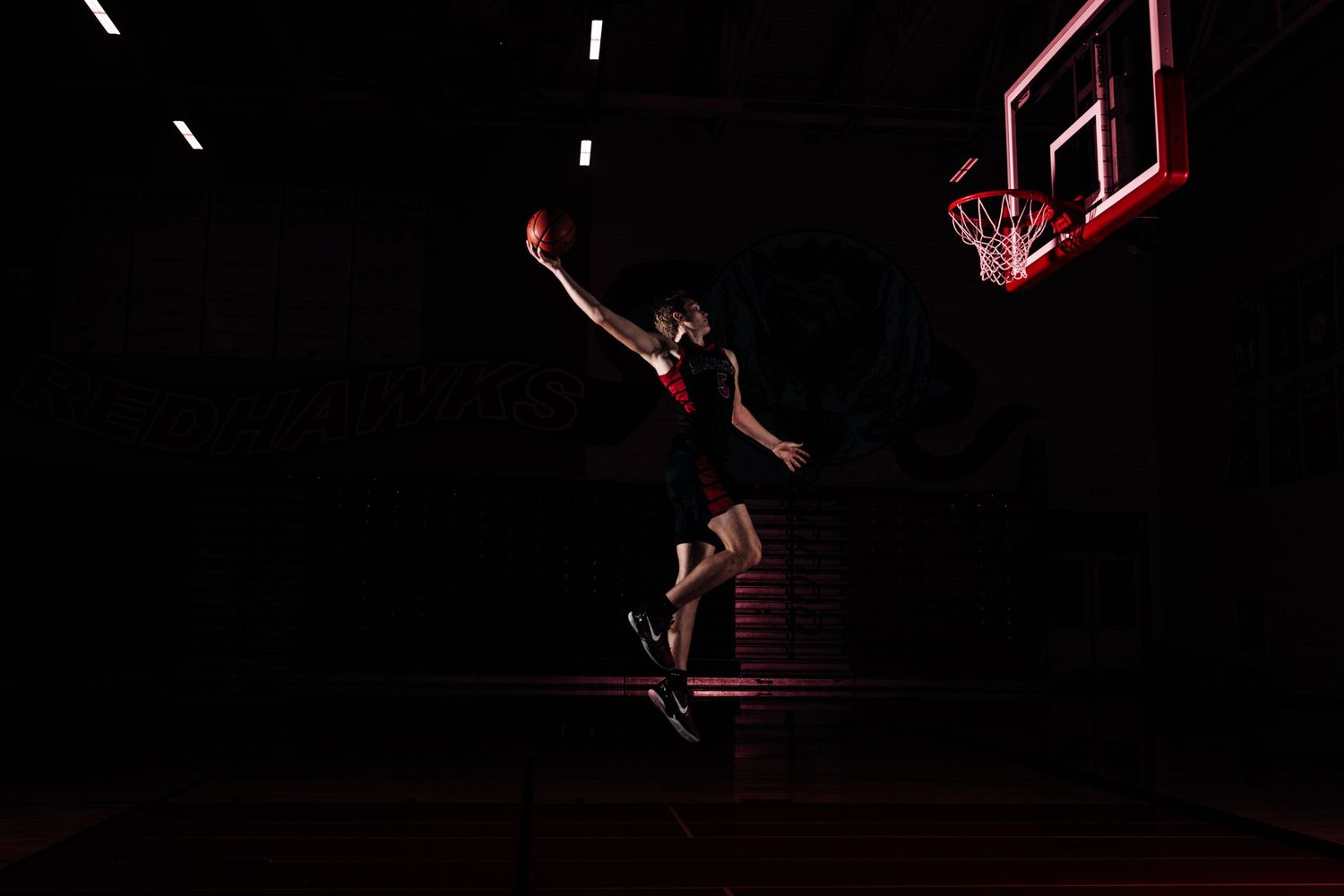 Basketball players soar through the dark arena performing a dramatic slam dunk beneath illuminated backboard.