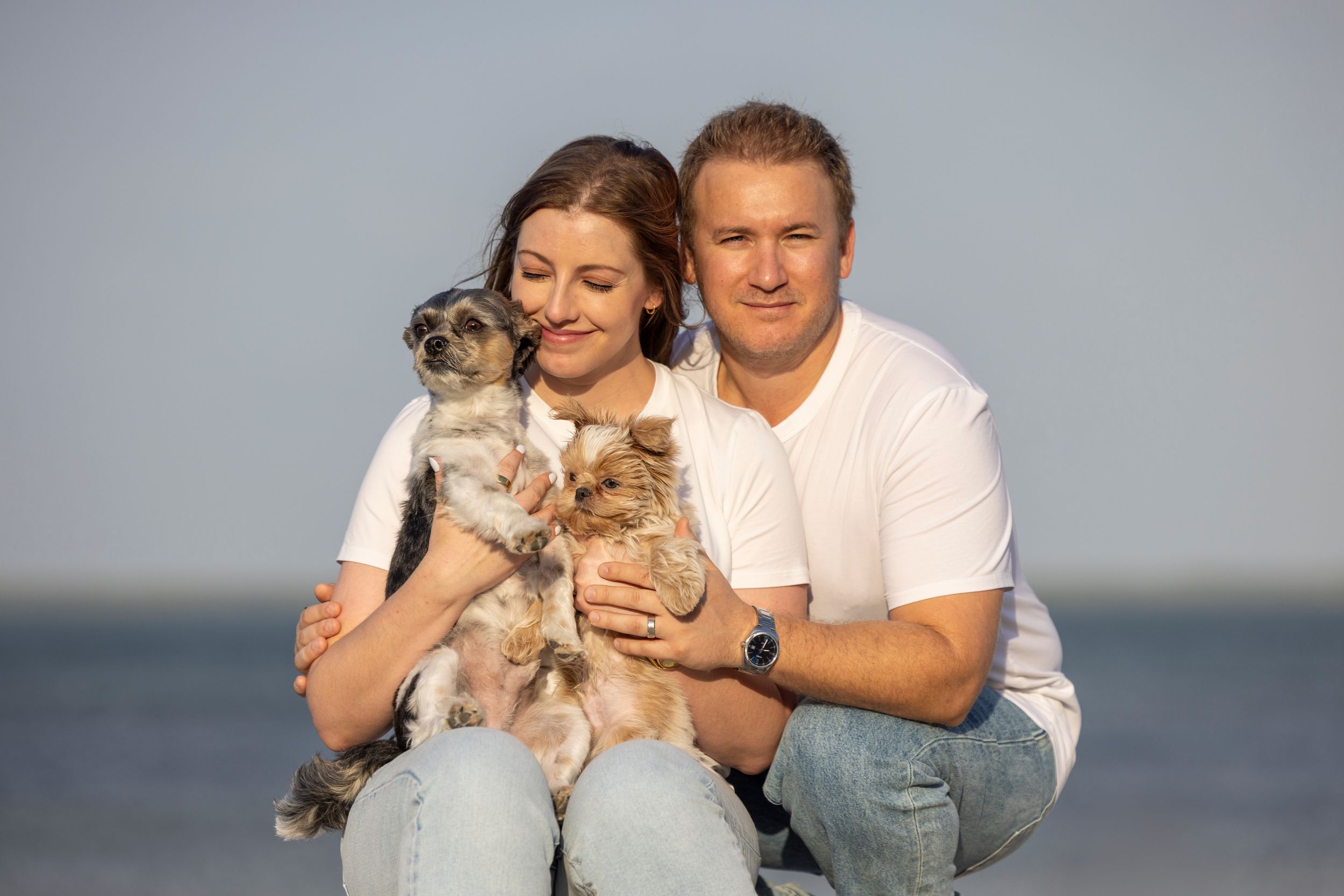 A smiling man and woman on a sandy beach, each cradling a small dog in their arms with the ocean in the background.