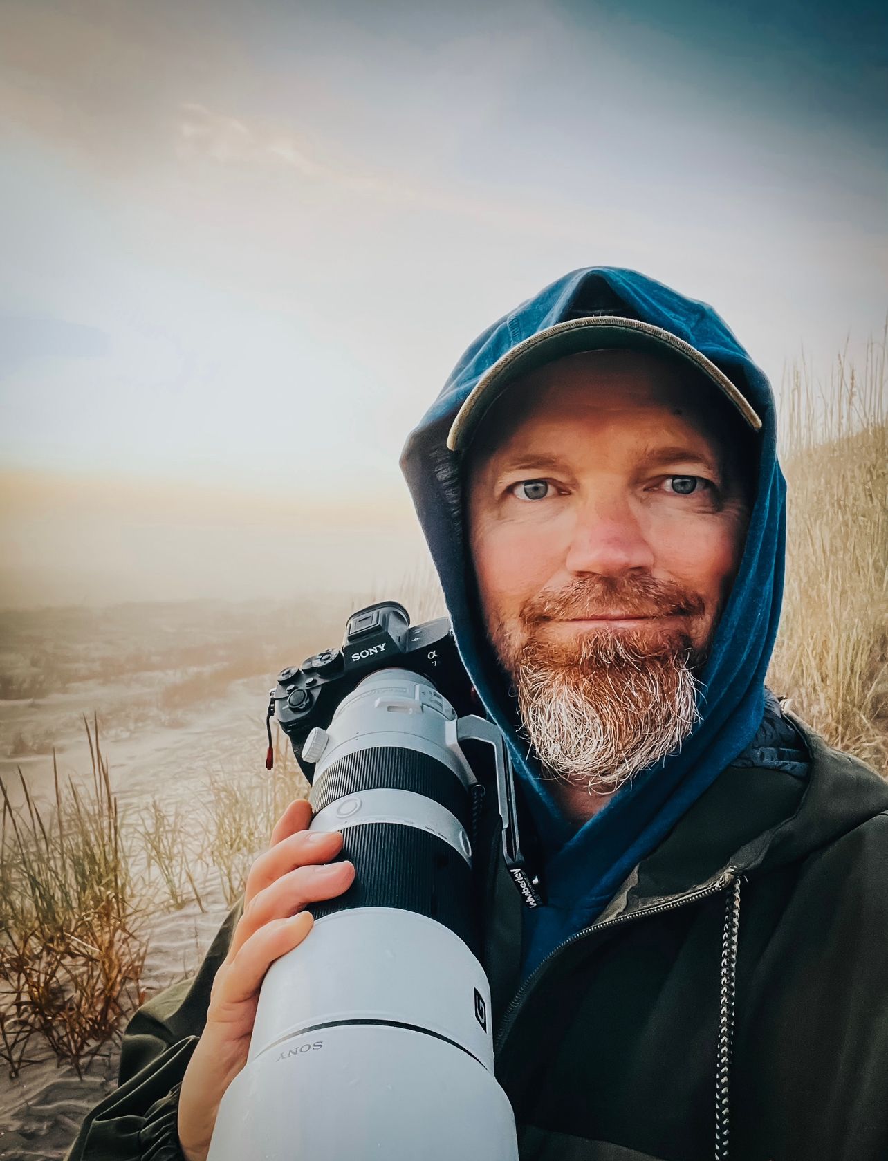 Wildlife photographer in a blue hooded jacket holding a large telephoto lens on a misty morning at the beach.