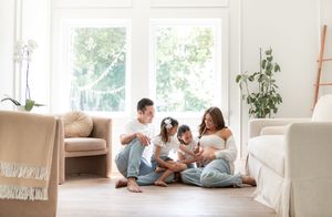 Family sitting on floor playing with dog in bright living room with white furniture.
