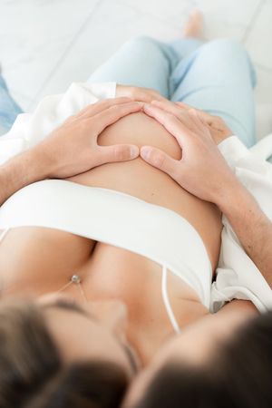 Close-up shot of hands resting on pregnant belly against light background.