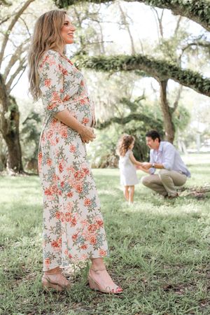 Expecting mother in floral dress poses in park with family playing in background.