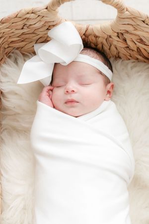 Sleeping newborn wrapped in white swaddle with bow headband on tan angel wing backdrop.
