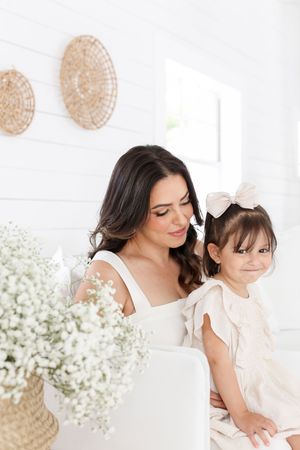 Mother and daughter in white dresses pose together with baby's breath flowers in bright room.