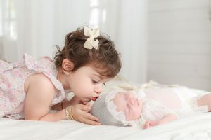 Young child in pink dress leaning over to look at something while lying on white bedding.