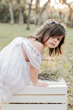 Dreamy portrait of someone in a white lace dress and flower crown leaning on a white box outdoors.