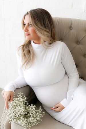 Maternity photo of woman in white dress sitting on beige chair with baby's breath flowers.