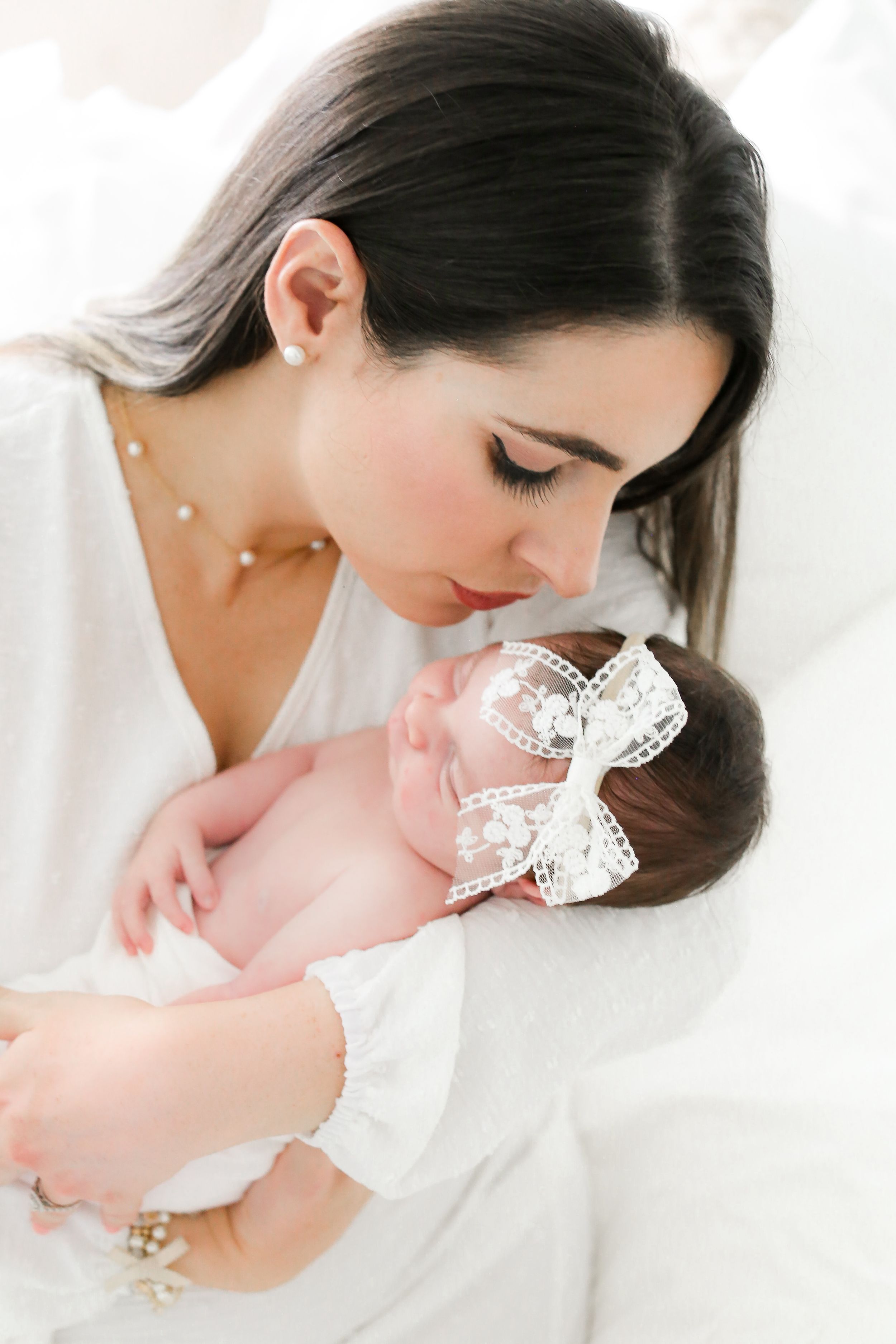 Tender moment between mother and newborn baby wearing lace bow in bright white setting.