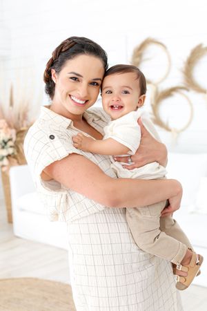 A tender moment captured as a woman in a white dress holds a smiling baby in a bright, airy setting.