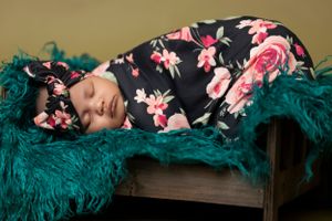 A sleeping newborn wrapped in a floral black and pink swaddle rests on a teal-colored furry blanket.