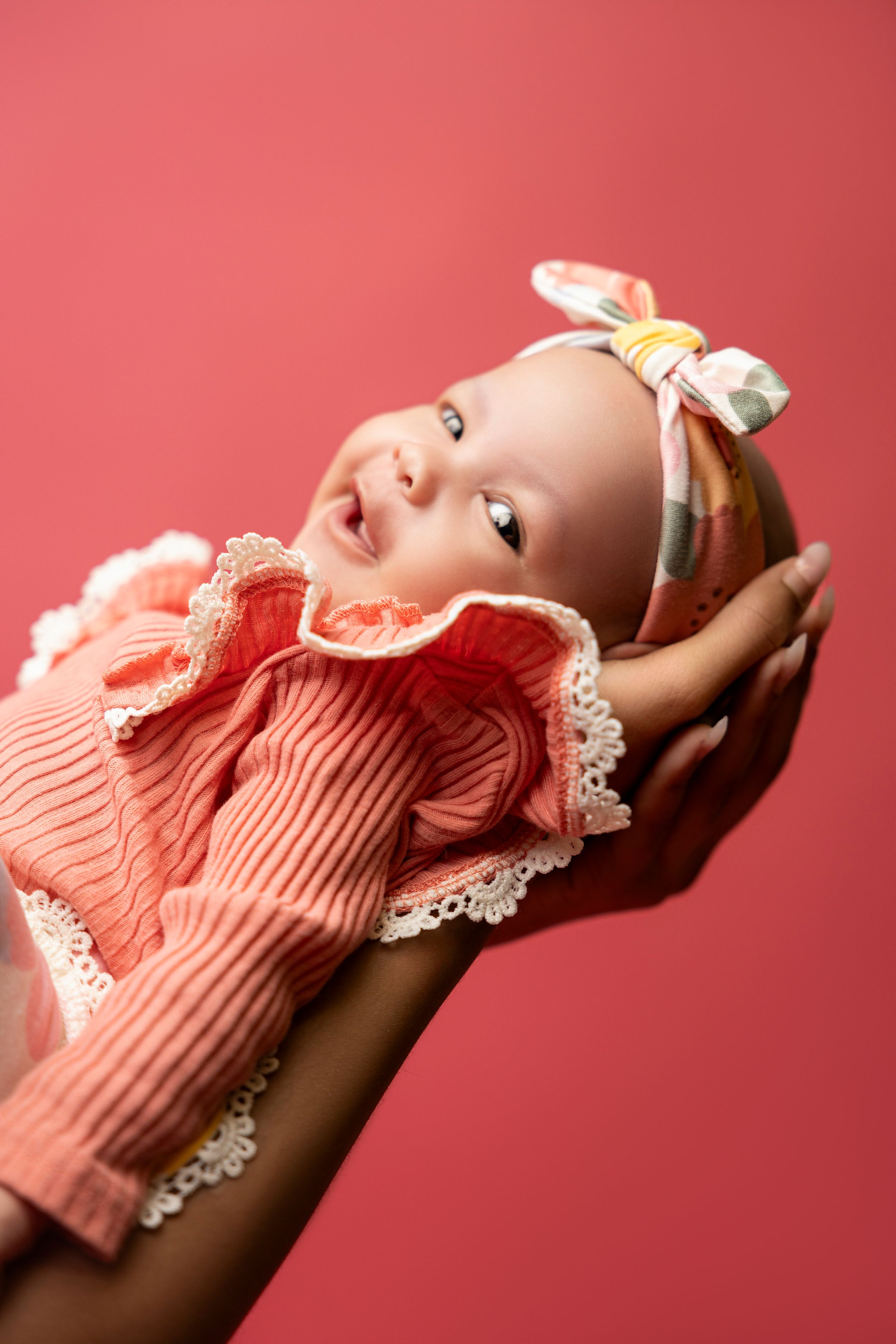 Small infant wearing a peachy-coral knit outfit with lace trim and headband lies against red background.
