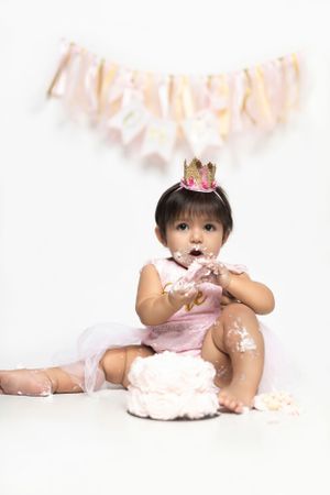 A baby wearing a pink outfit and tiny crown sits on floor with a smashed birthday cake during a cake smash photo session.