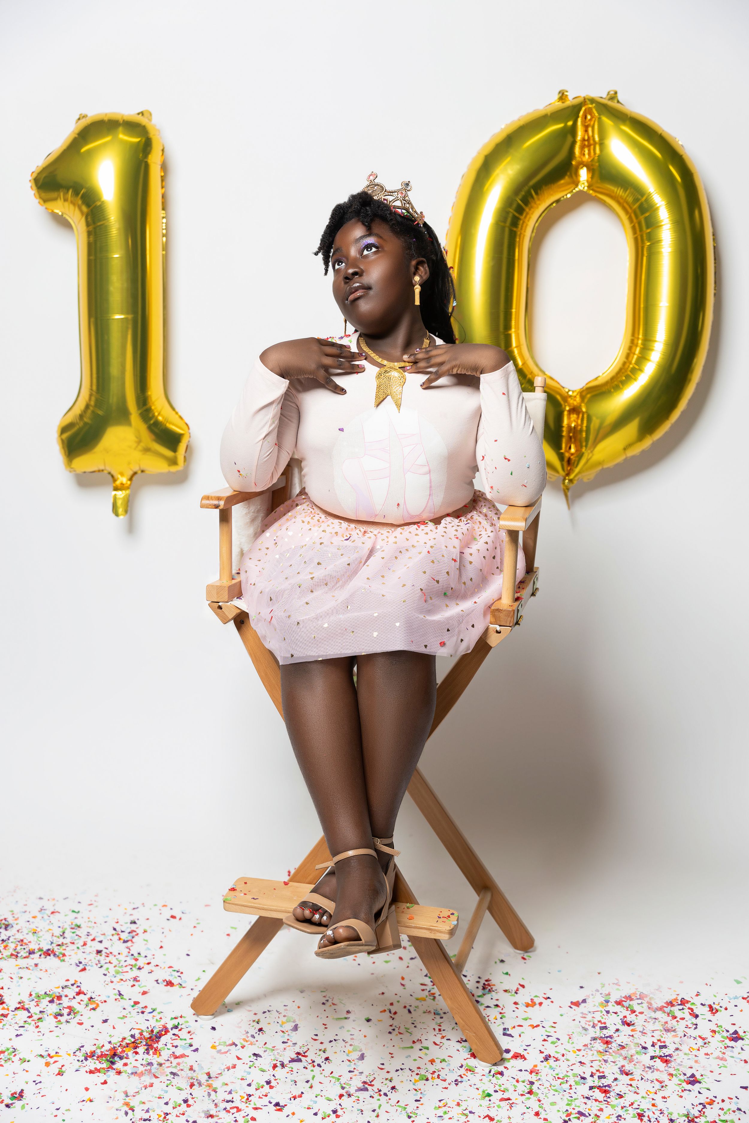 Birthday celebration with gold number 10 balloons, featuring person in pink dress seated in director's chair with confetti.