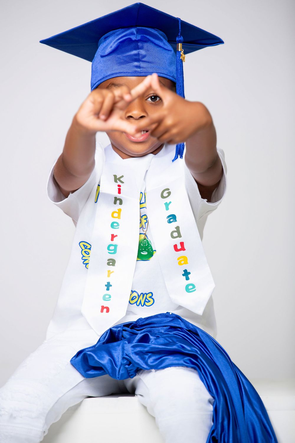 A graduate in royal blue cap and gown and decorated sash makes a playful hand gesture during a portrait session.