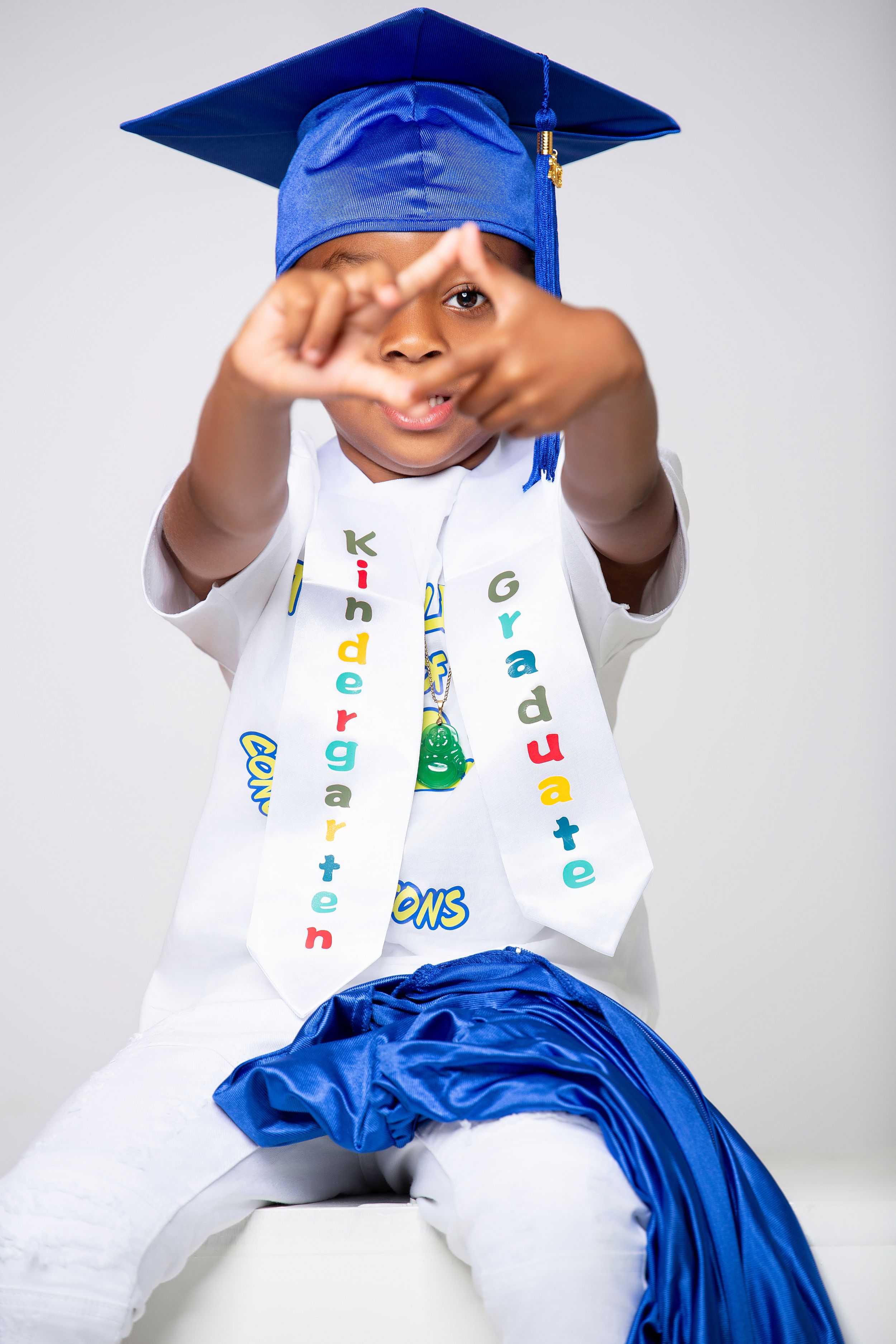 A graduate in royal blue cap and gown and decorated sash makes a playful hand gesture during a portrait session.