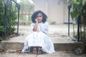 A small child in a white lace dress sits thoughtfully on garden steps with brown boots.