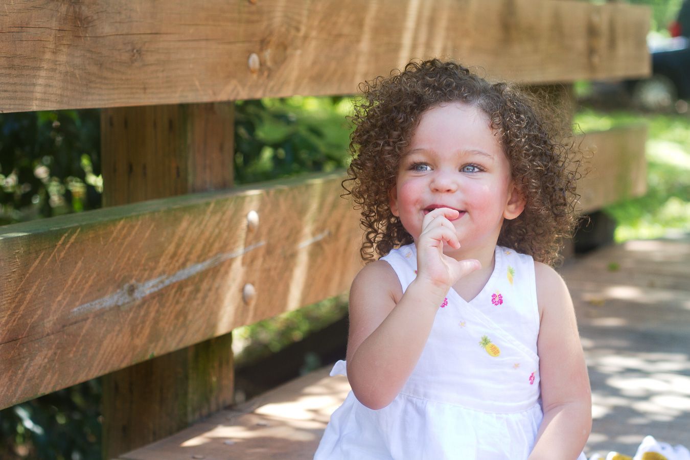Young child with curly hair wearing a white summer dress poses playfully near a wooden fence on a sunny day.