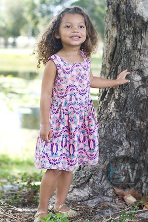 A child in a pink and blue patterned summer dress stands next to a large tree trunk near a pond on a sunny day.