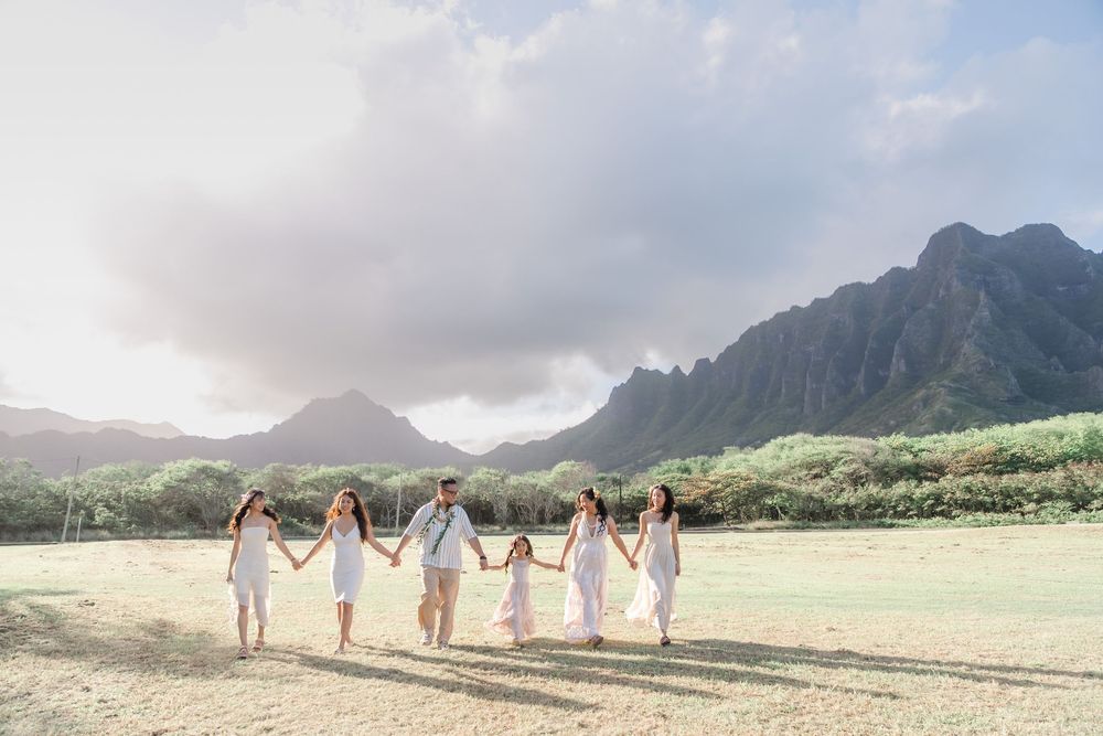 A group in white dresses walks through a scenic Hawaiian valley with dramatic mountain peaks in the background.