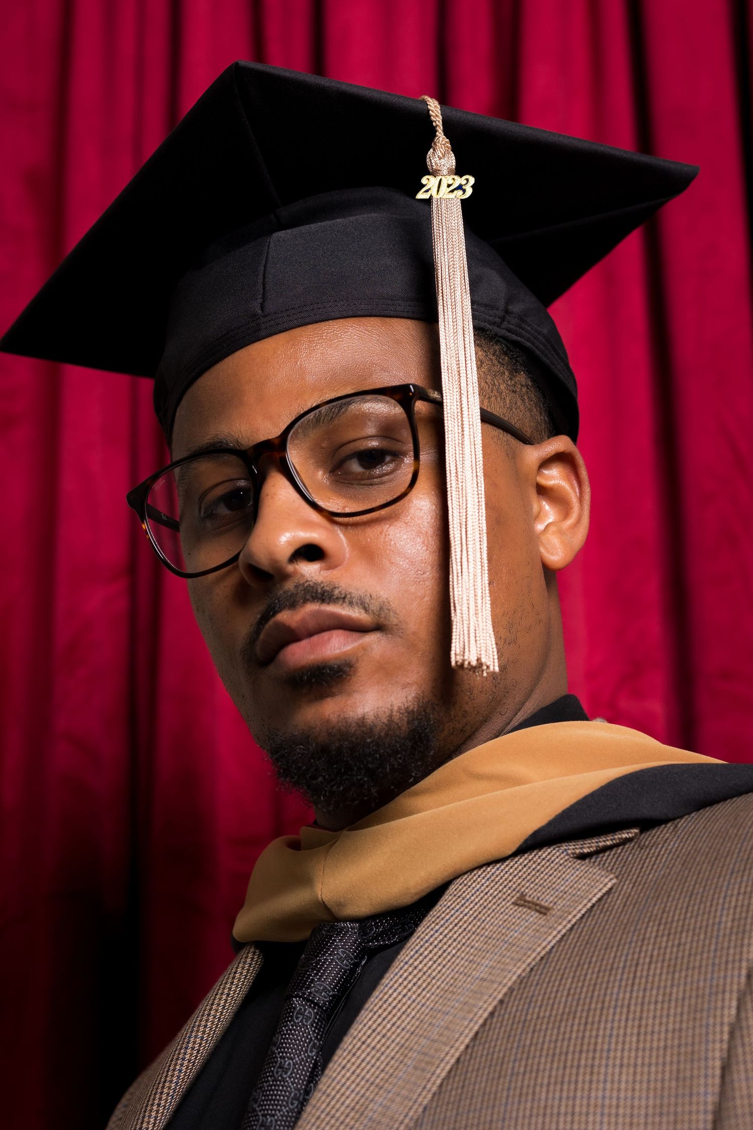 Graduate in cap and gown poses against red curtain backdrop.