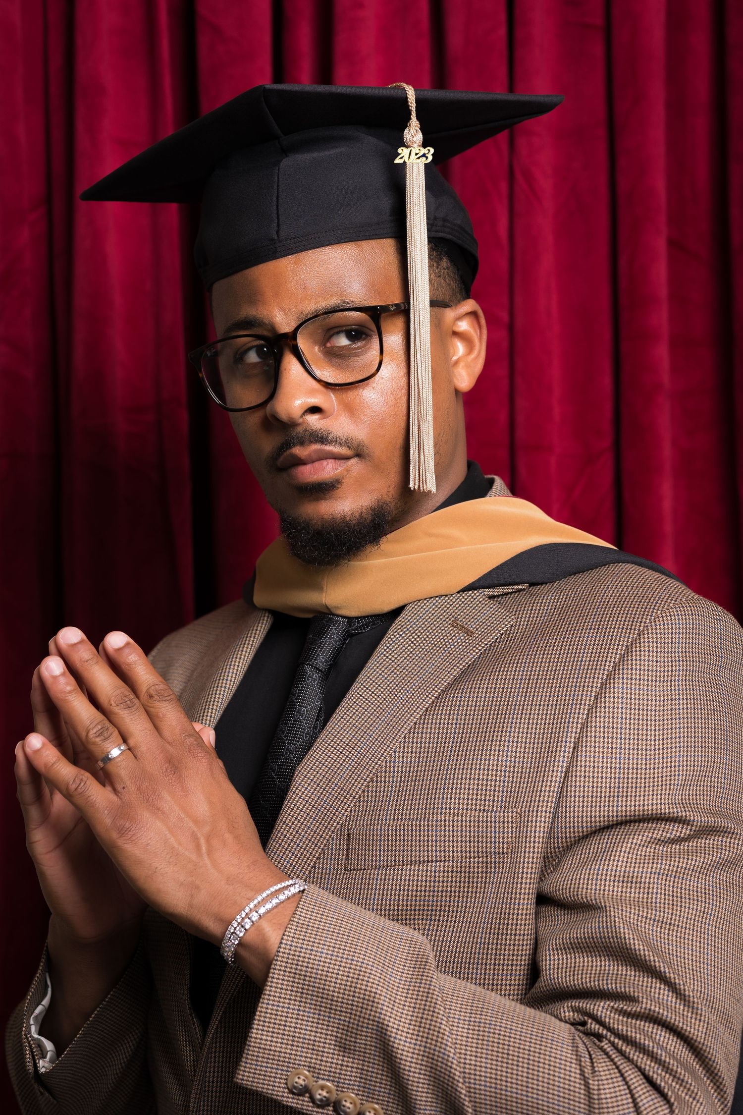 Graduate in brown suit and cap poses with prayer hands against red backdrop.