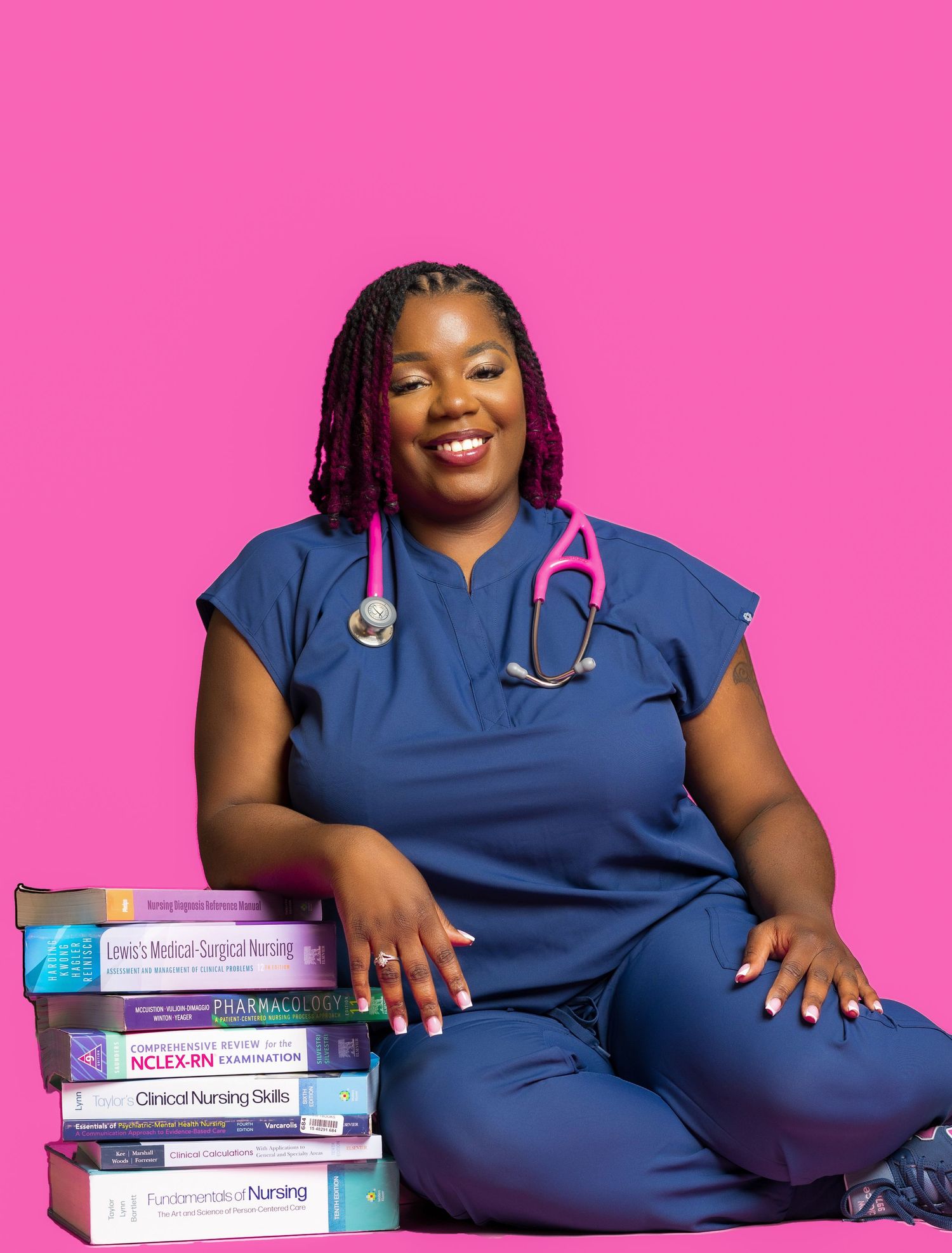 Healthcare professional in blue scrubs poses with medical textbooks against pink background.