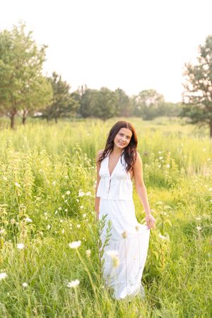 Kylie in a flowing white dress walks through a sunlit meadow filled with wildflowers and tall grass for her senior photos in Des Moines.