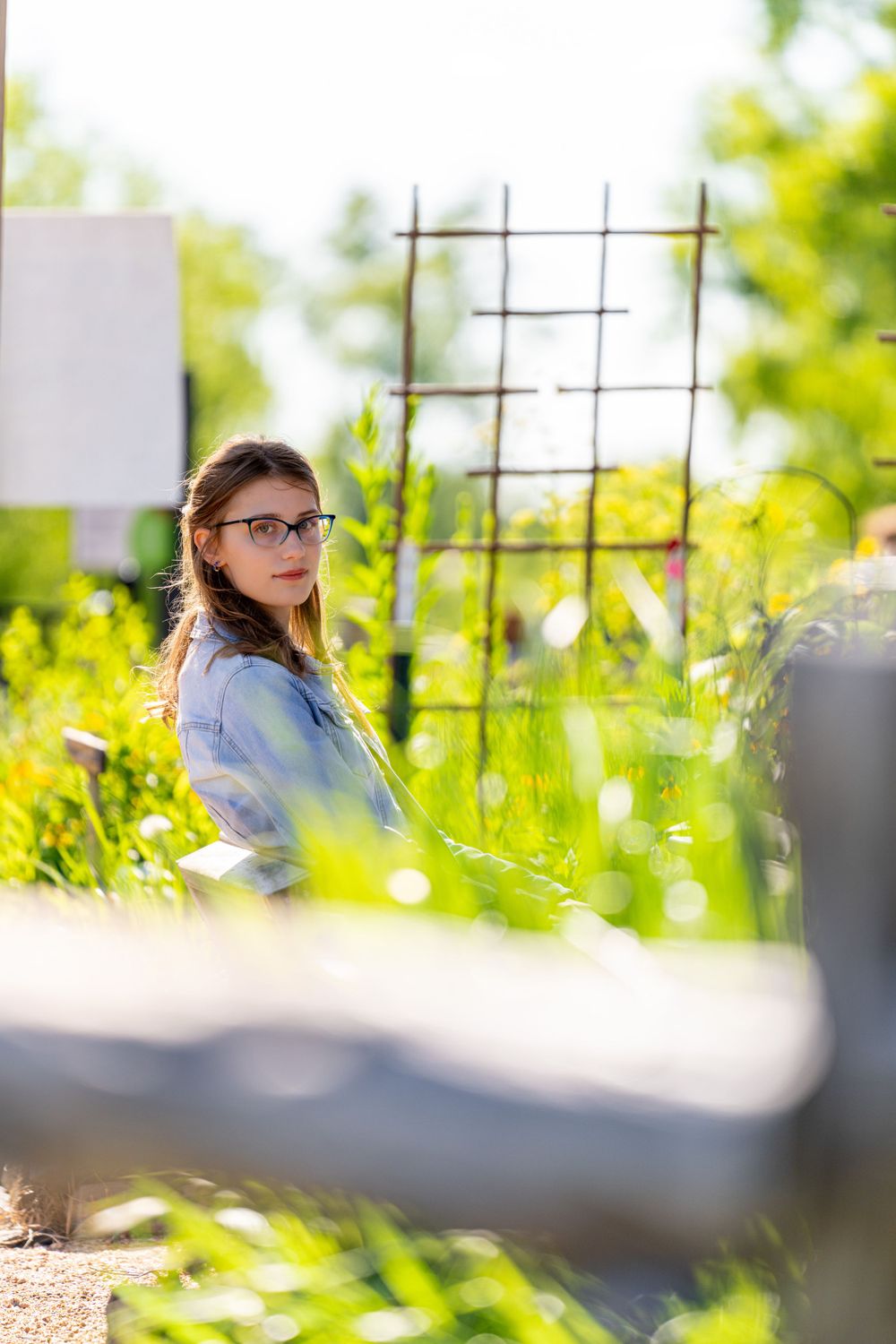 Student in denim jacket sits among yellow wildflowers next to garden trellis.