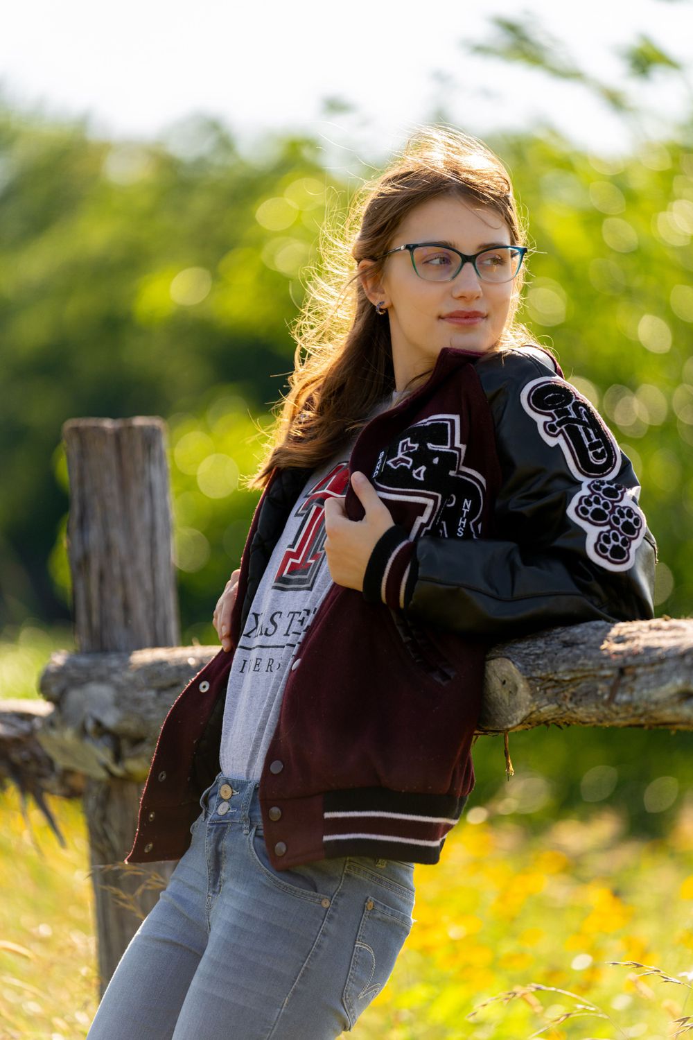 Student in varsity jacket poses against wooden fence with sunny yellow field background.