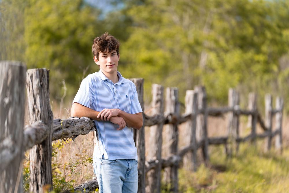 A teenage boy in a light blue polo shirt leans on a rustic wooden fence outdoors on a sunny day.