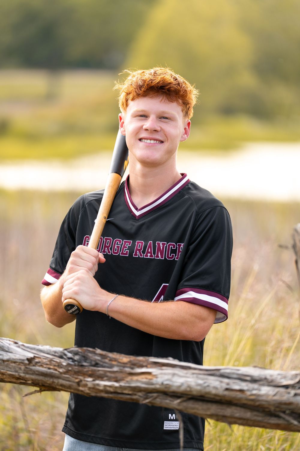 Smiling teen with red hair holds baseball bat wearing George Ranch jersey number 4 outdoors near wooden fence.